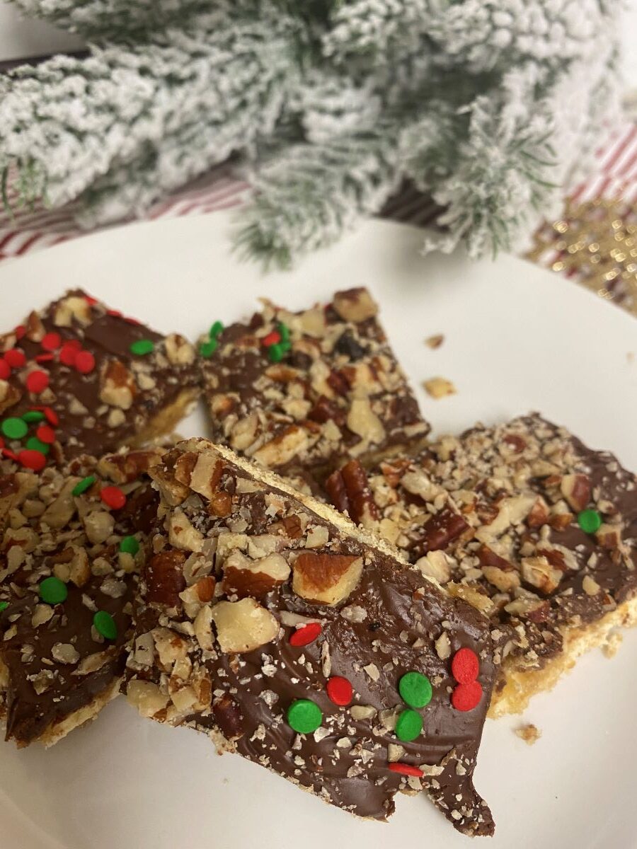Pile of saltine toffee on white plate with evergreen branch in background.