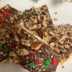 Saltine toffee in pile in white plate with Christmas tree branches in background.