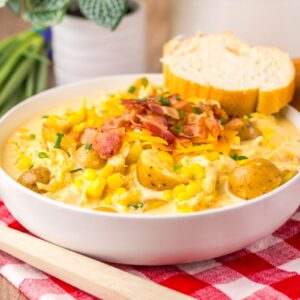 White bowl of chicken corn chowder with slice of French bread on the side on red gingham napkin.