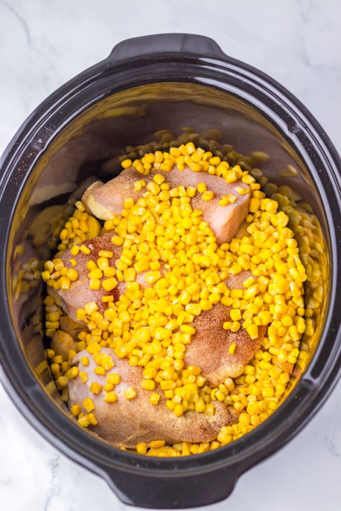 Raw chicken and corn in black slow cooker.