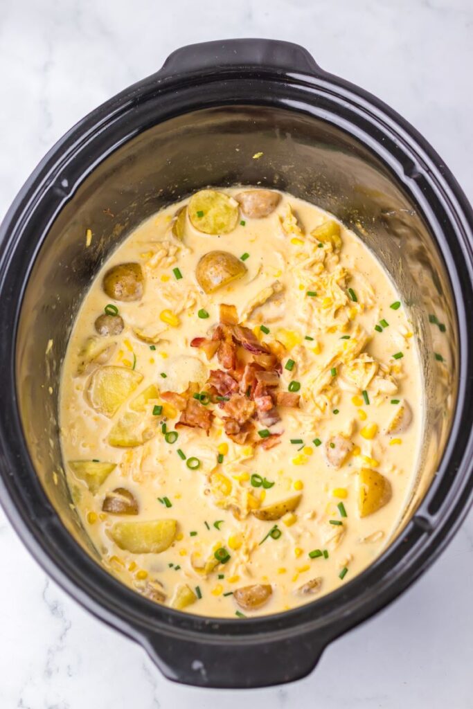 Mixture of corn, potatoes, and cream with bacon and scallions on top in slow cooker.
