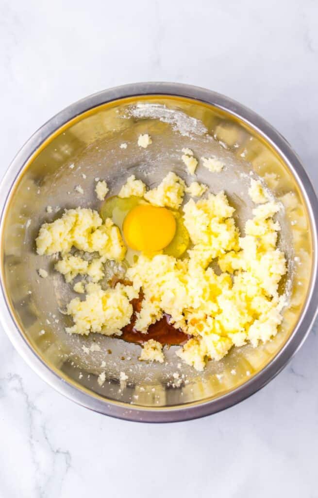 A metal mixing bowl with creamed butter and sugar, an egg, and vanilla extract on top, ready to be mixed together.