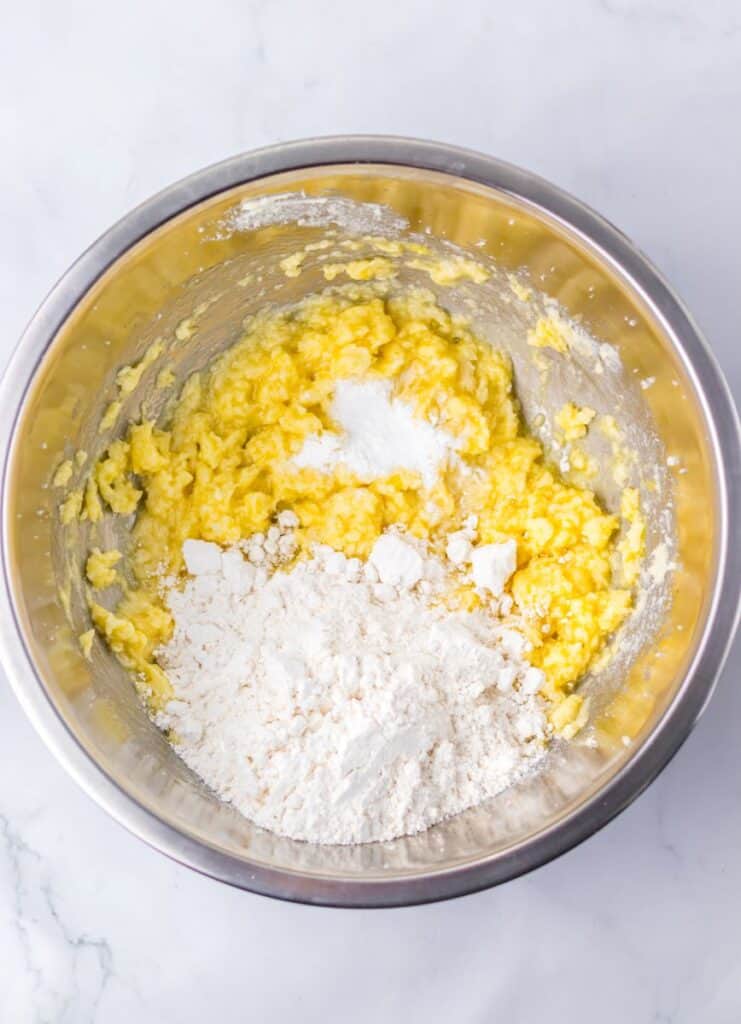 A metal bowl containing creamed yellow mixture with sugar and flour on top, ready to be mixed together for baking, sits on a white surface.