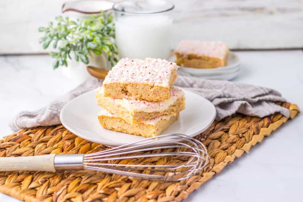 Three frosted sugar cookie bars stacked on a white plate sit on a woven placemat with a whisk, plant, glass of milk, and more cookie bars on a plate in the background.