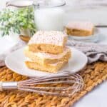 Three frosted sugar cookie bars stacked on a white plate sit on a woven placemat with a whisk, plant, glass of milk, and more cookie bars on a plate in the background.