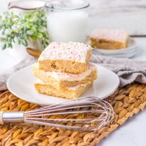 Three frosted sugar cookie bars stacked on a white plate sit on a woven placemat with a whisk, plant, glass of milk, and more cookie bars on a plate in the background.