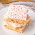 Close up of three light brown cookie bars with red and white speckled frosting stacked on white plate.