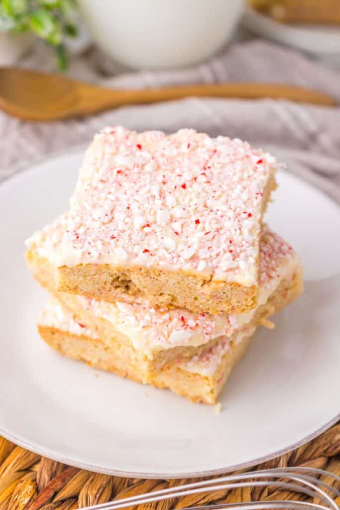 Close up of three light brown cookie bars with red and white speckled frosting stacked on white plate.