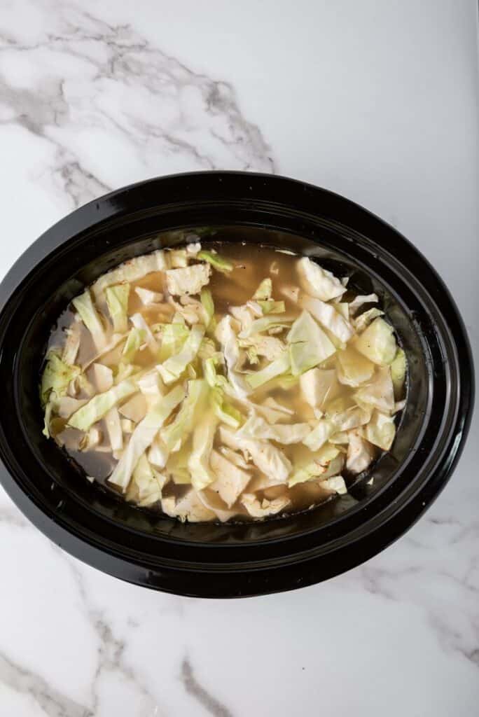 Top view of black slow cooker with chicken stock and green cabbage inside.