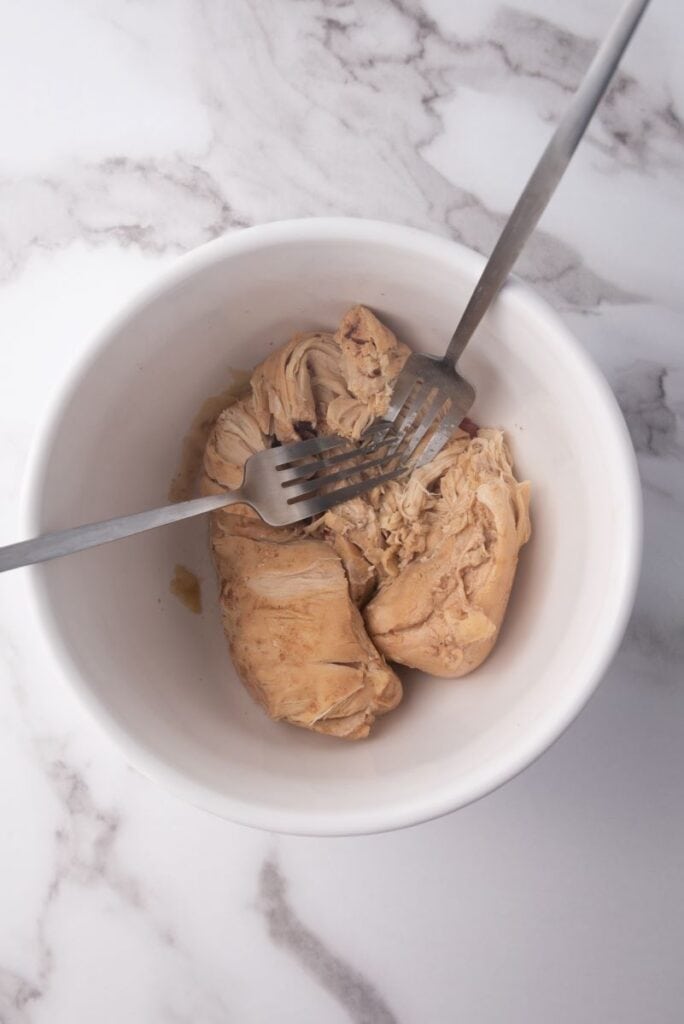 Cooked chicken breast in white bowl with two silver forks.