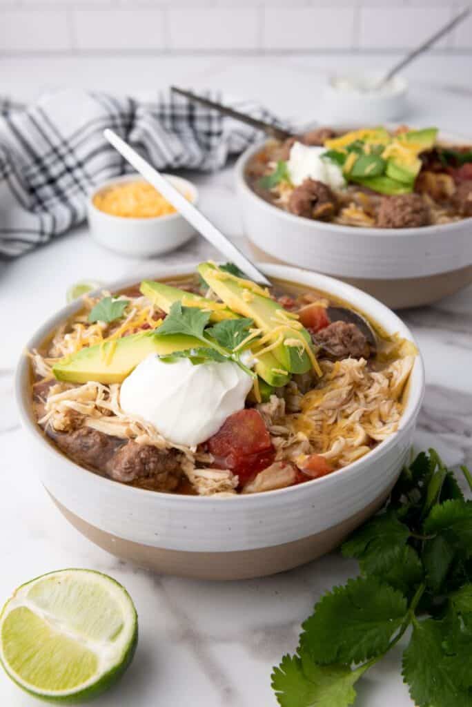 Close up of two white bowls of taco soup topped with shredded cheese, cilantro, and sliced avocados. Each bowl has a silver spoon. Bowls are on white marble counter with sliced limes and a bunch of cilantro. Small bowl of yellow shredded cheese in background.