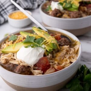 Top view of two white bowls of taco soup topped with shredded cheese, cilantro, and sliced avocados. Each bowl has a silver spoon. Bowls are on white marble counter with a bunch of cilantro. Small bowl of yellow shredded cheese in background.