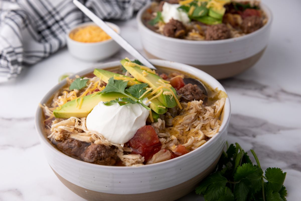 Top view of two white bowls of taco soup topped with shredded cheese, cilantro, and sliced avocados. Each bowl has a silver spoon. Bowls are on white marble counter with a bunch of cilantro. Small bowl of yellow shredded cheese in background.