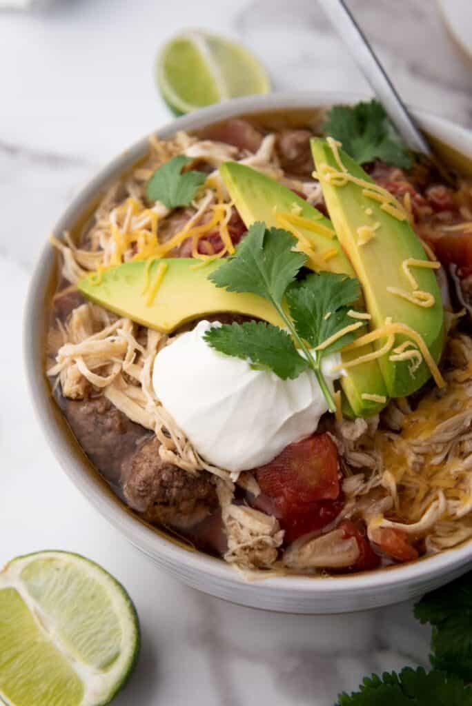 Close up of white bowl of taco soup topped with shredded cheese, cilantro, and sliced avocados. Bowl has a silver spoon. Bowl is on white marble counter with sliced limes and a bunch of cilantro.