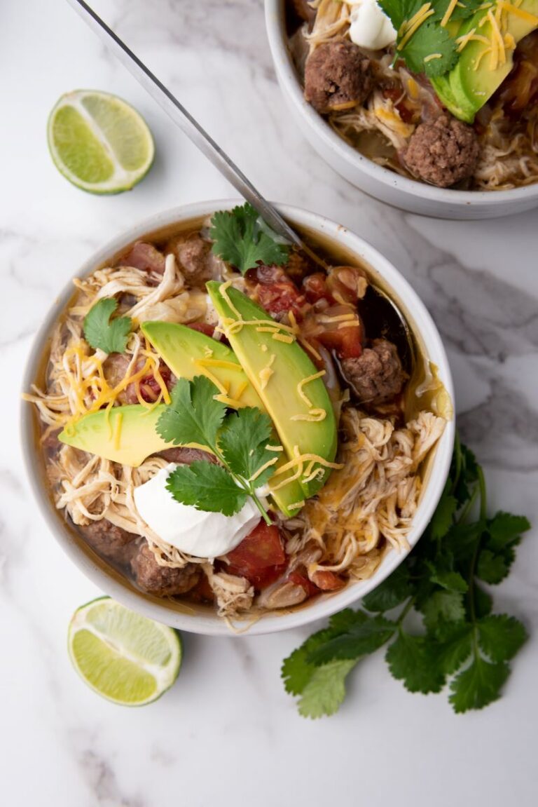 Top view of two white bowls of taco soup topped with shredded cheese, cilantro, and sliced avocados. Each bowl has a silver spoon. Bowls are on white marble counter with sliced limes and a bunch of cilantro.