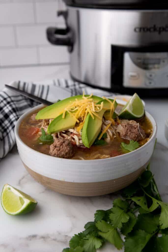 White bowl of taco soup topped with shredded cheese, cilantro, and sliced avocados. Bowl has a silver spoon. Bowl is on white marble counter with sliced limes and a bunch of cilantro. Small bowl of yellow shredded cheese, black gingham towel and silver slow cooker in background.