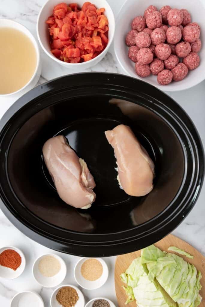 Top view of raw chicken breasts in black slow cooker. Bowls of other ingredients surrounding it.