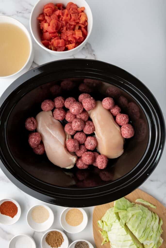 Top view of raw chicken breasts and raw meatballs in black slow cooker. Other ingredients in white bowls surrounding slow cooker.