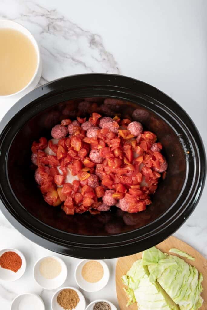 Roasted tomatoes on top of meatballs and chicken breasts in black slow cooker. Other ingredients in white bowls surrounding slow cooker.