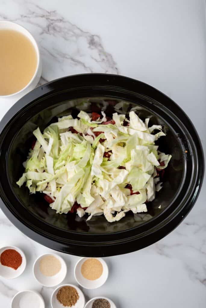 Green cabbage on top of tomatoes and meat in black slow cooker. Other ingredients in white bowls surrounding slow cooker.