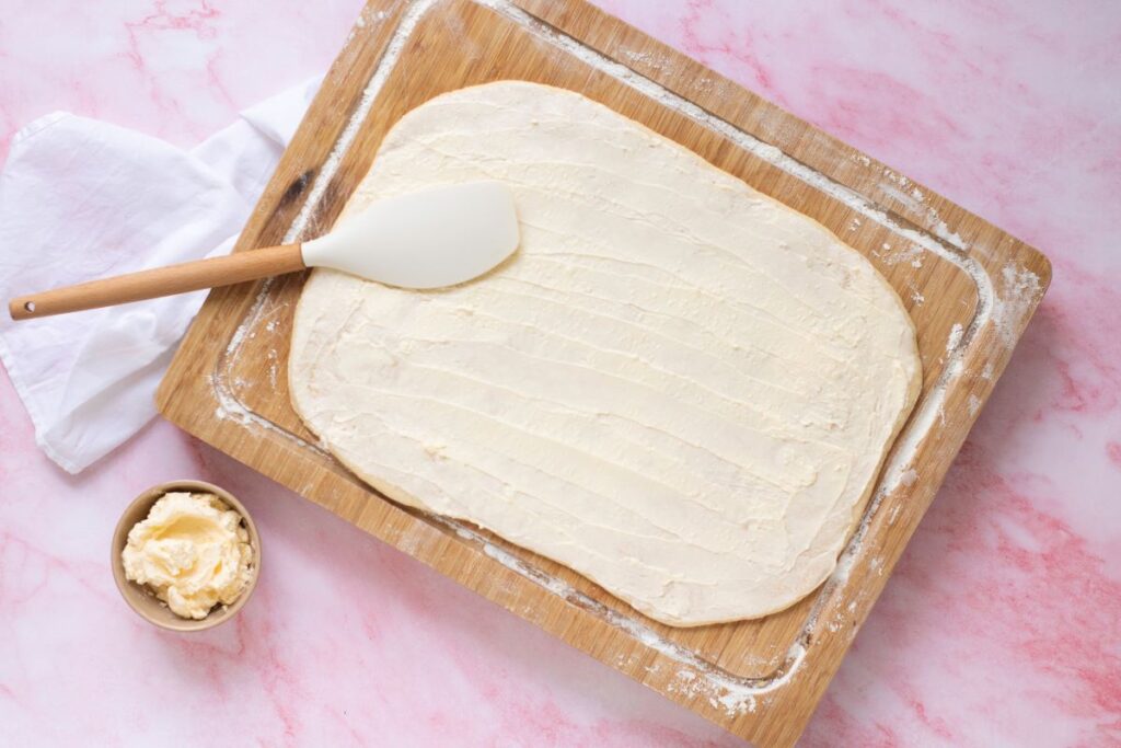 Flattened croissant dough with butter spread on top on wooden cutting board.