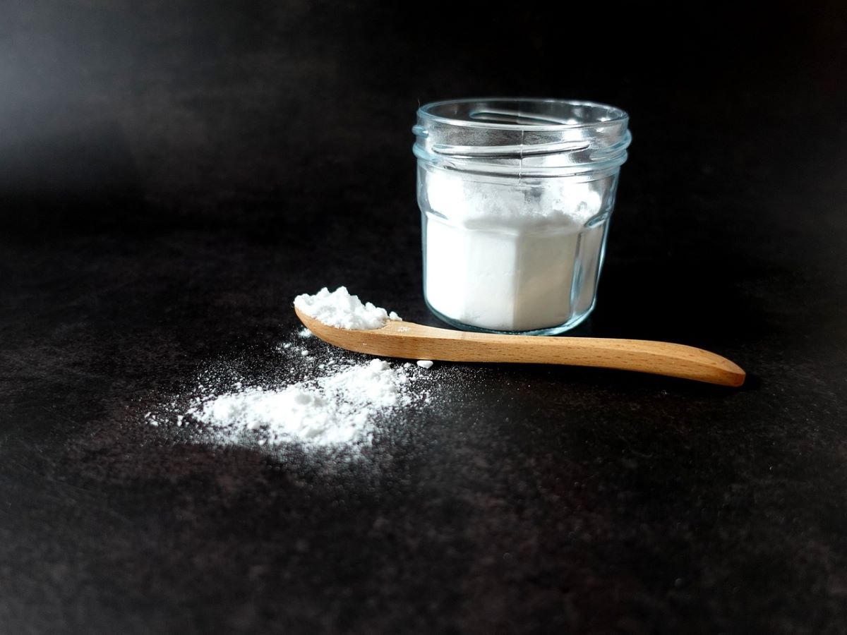 Wooden spoon with baking soda laying in front of glass jar full of baking soda.