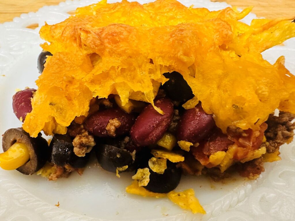 Side view of casserole with melted cheese, red kidney beans, corn, olives, and hamburger on white plate.