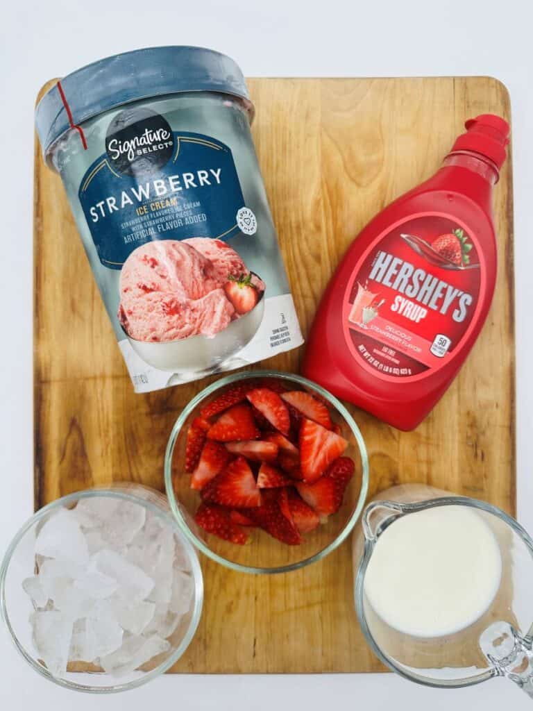 Ingredients for strawberry milkshake in glass bowls on wooden board.