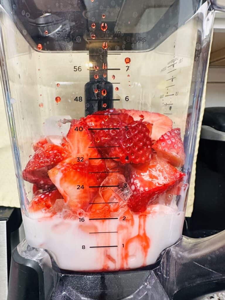 Milk, red syrup, strawberries, and ice in clear blender.