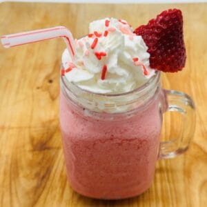 Pink drink in glass jar with handle topped with whipped cream and red sprinkles and a sliced strawberry with bent red and white straw.