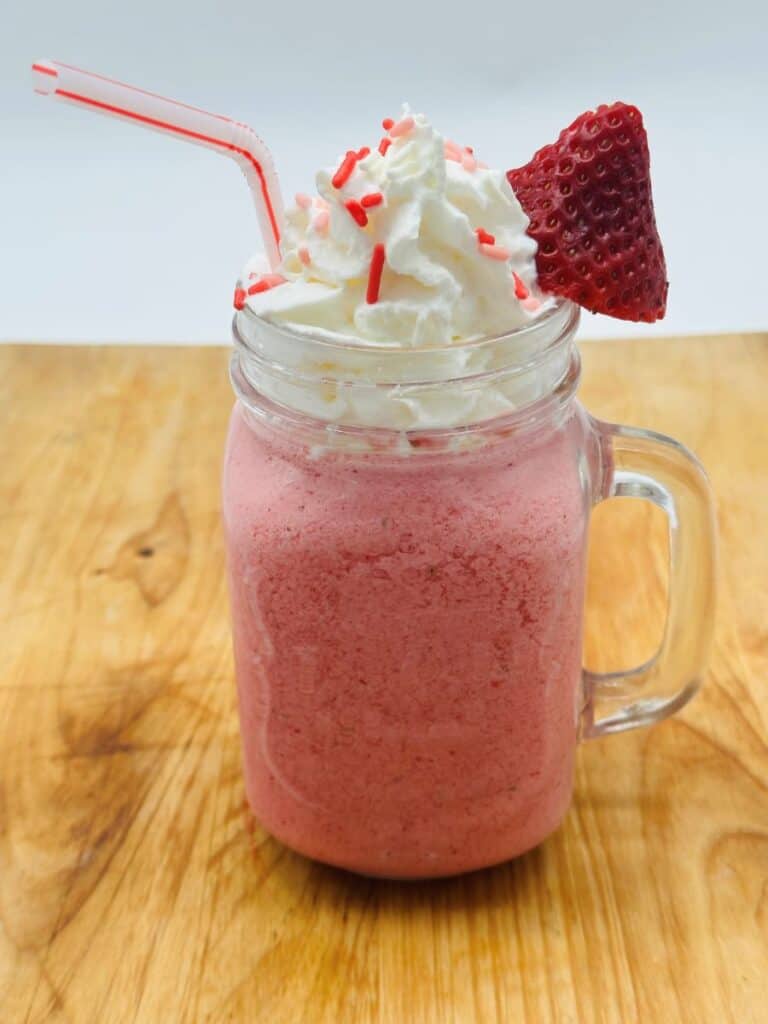 Pink drink in glass jar with handle topped with whipped cream and red sprinkles and a sliced strawberry with bent red and white straw.