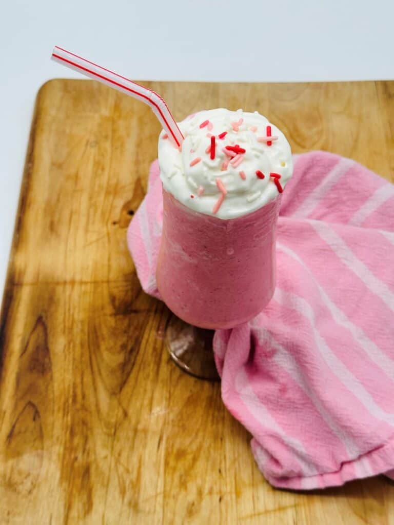 Pink drink in stemmed glass topped with whipped cream and red sprinkles with bent red and white straw.