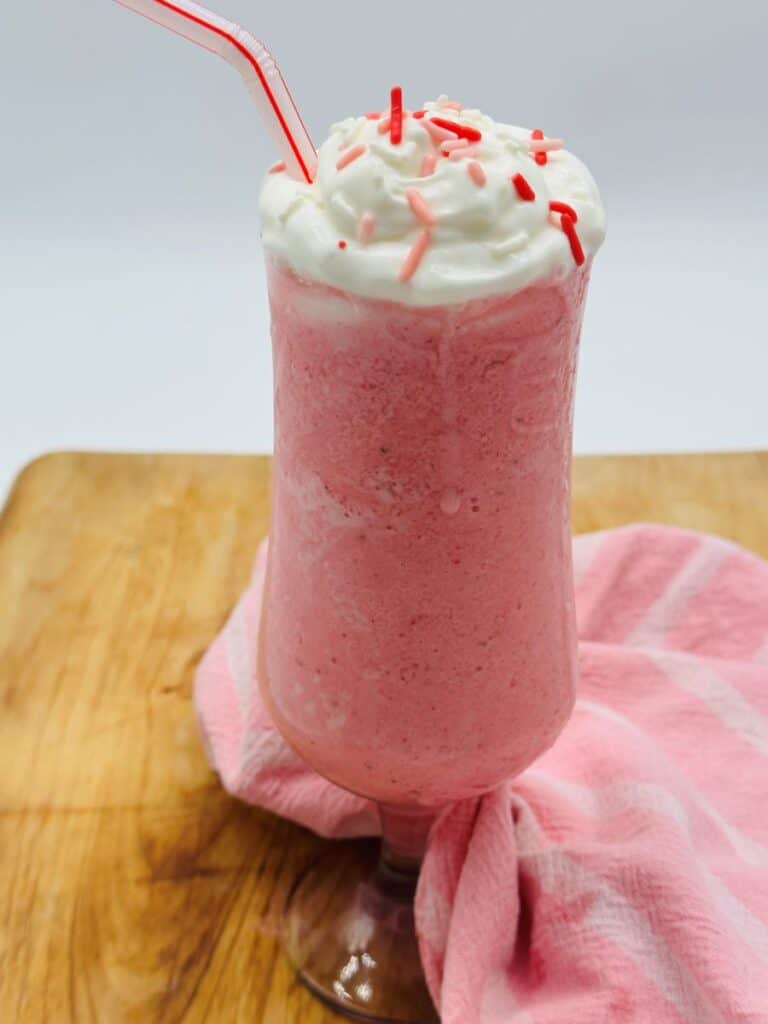 Pink drink in stemmed glass topped with whipped cream and red sprinkles and a sliced strawberry with bent red and white straw.