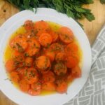 Sliced carrots in orange sauce on white plate. Greenery on wooden board above. Grey and white towel below.