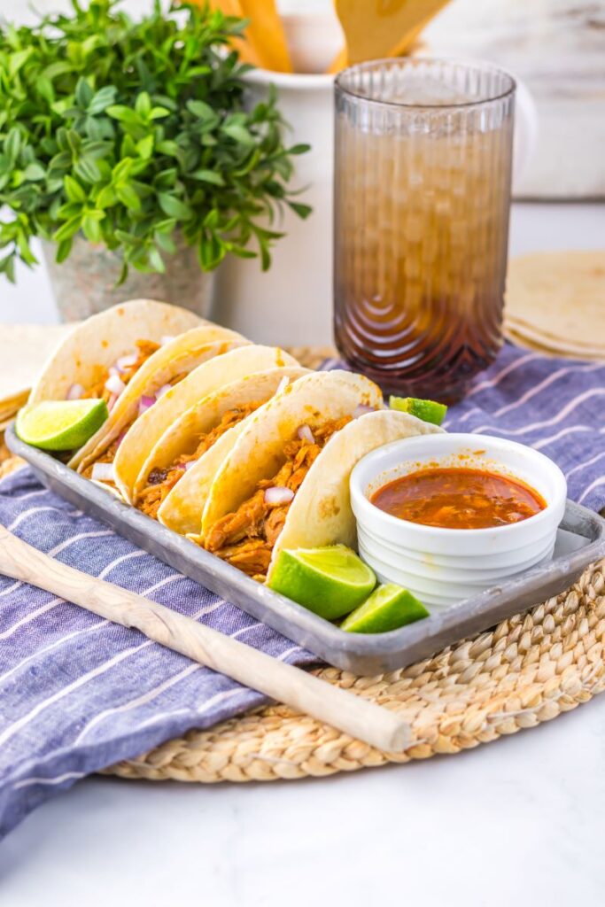 Flour tortillas with shredded pork on pewter tray with limes and white jar of red sauce.