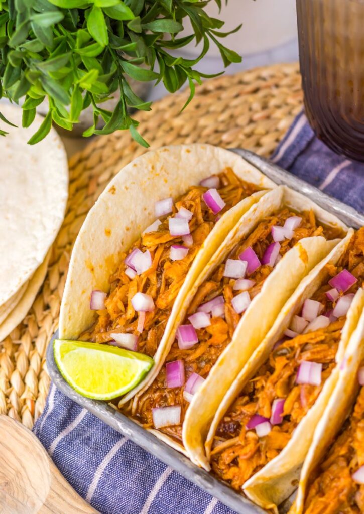 Close up of flour tortillas with shredded pork on pewter tray with limes and white jar of red sauce.