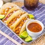 Flour tortillas with shredded pork on pewter tray with limes and white jar of red sauce.