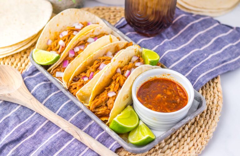 Flour tortillas with shredded pork on pewter tray with limes and white jar of red sauce.