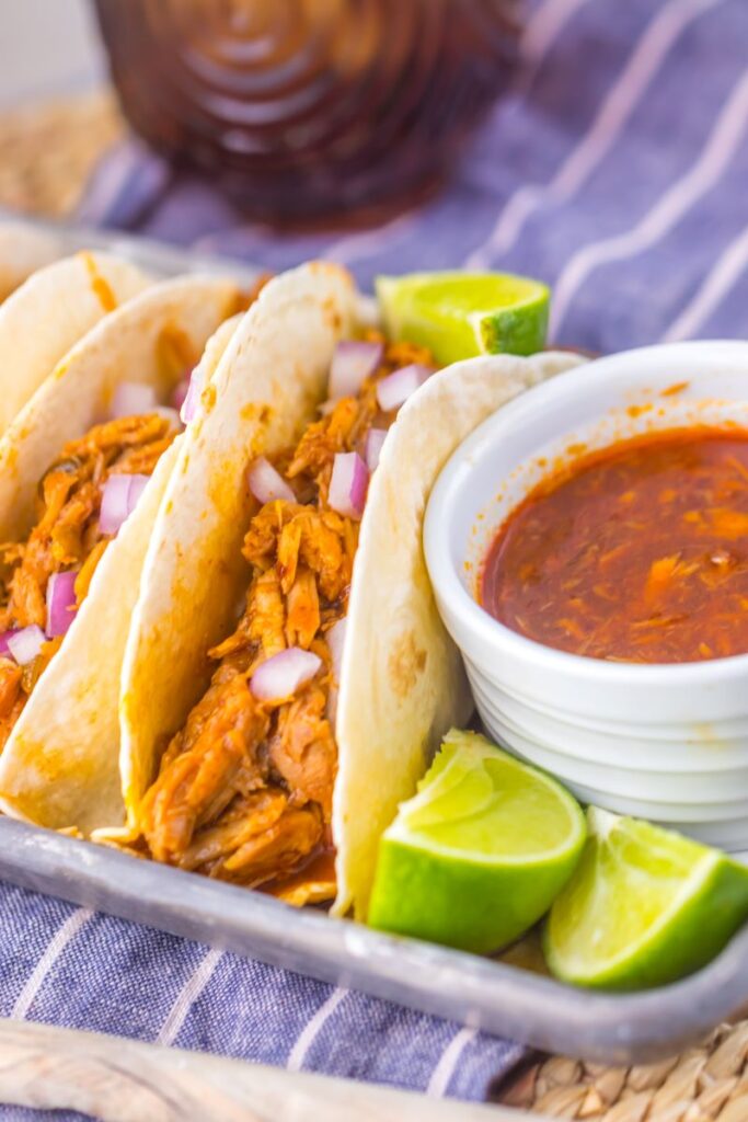 Close up of flour tortillas with shredded pork on pewter tray with limes and white jar of red sauce.