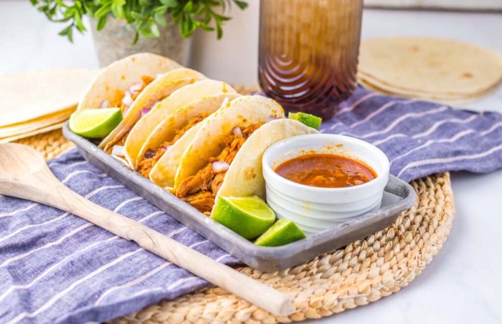 Flour tortillas with shredded pork on pewter tray with limes and white jar of red sauce.