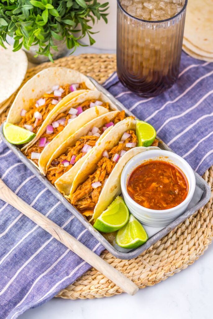 Flour tortillas with shredded pork on pewter tray with limes and white jar of red sauce.