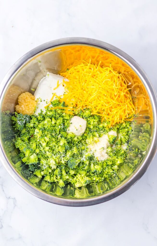 Chopped broccoli, shredded cheddar cheese, cream cheese, butter, parmesan cheese, and spices in silver bowl.