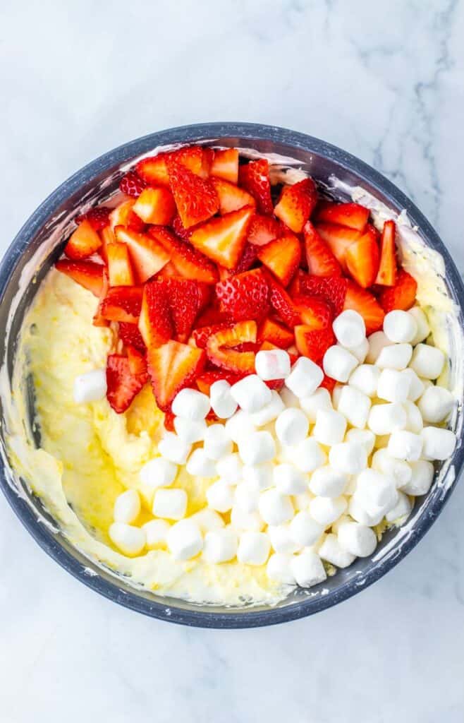 Top view of mini marshmallows and chopped strawberries on yellow mixture in blue bowl.