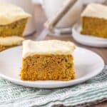 Side view of zucchini cake with white frosting on white plate.