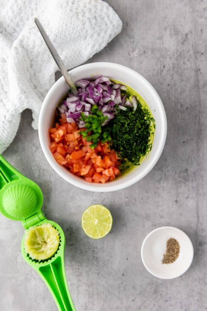 Top view of white bowl of chopped ingredients with chopped lime and small white bowl of pepper in front.