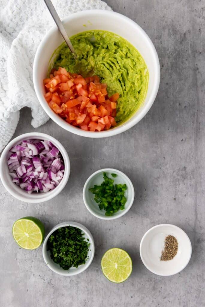 White bowl with green guacamole and diced tomatoes. Small white dishes with diced red onions, diced jalapenos, diced cilantro, and black pepper in front.