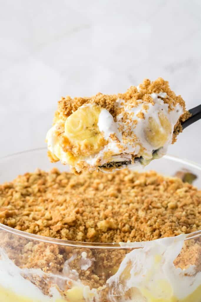 Spoon lifting one serving of layered pudding over clear glass trifle bowl.