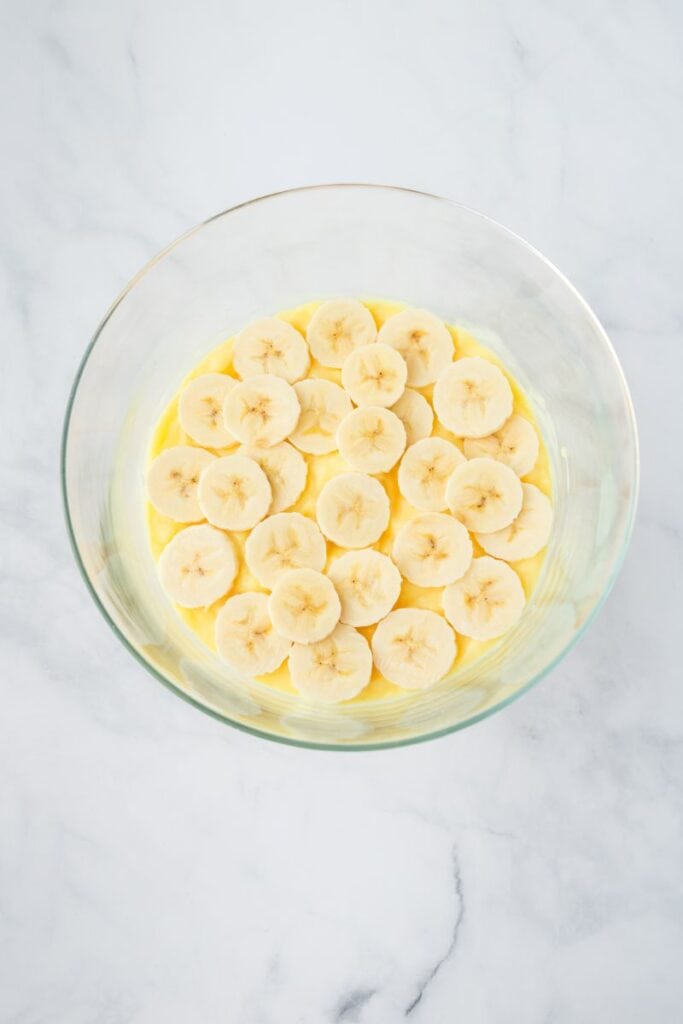 Overhead view of sliced bananas on top of yellow pudding in clear glass bowl.