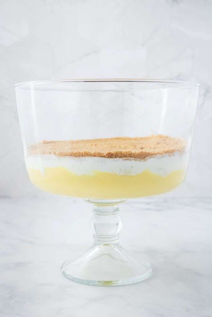 Side view of clear glass trifle bowl with one layer of yellow pudding, one layer of whipped cream, and one layer of crushed cookies.