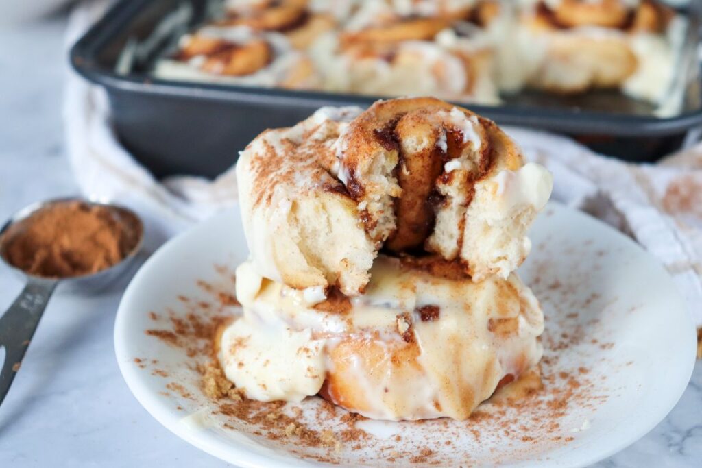 Close up of two stacked cinnamon rolls, the top one with a bite out of it, on a white plate in front of a square metal pan of cinnamon rolls.